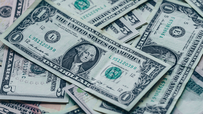 A close-up view of several scattered U.S. one-dollar bills, showing the front side with George Washington’s portrait and various serial numbers and seals visible.