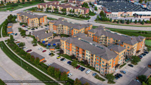Aerial view of a large, modern apartment complex with multiple colorful buildings, green lawns, and parking lots, surrounded by roads and nearby commercial buildings in a suburban area.