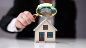 A person in a suit holds a magnifying glass over a small model house, suggesting close inspection or evaluation of real estate.