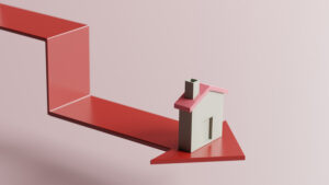 A small house model sits on a red downward-pointing arrow, symbolizing a decline, against a pale pink background. The image visually conveys the concept of falling real estate or housing market values.