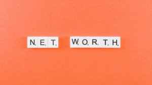 Scrabble letter tiles on an orange background spell out NET WORTH.