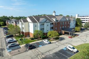 Aerial view of a multi-story apartment building with red brick and light siding, surrounded by trees. Several cars are parked in adjacent parking lots with visible handicap spaces. Blue sky above.