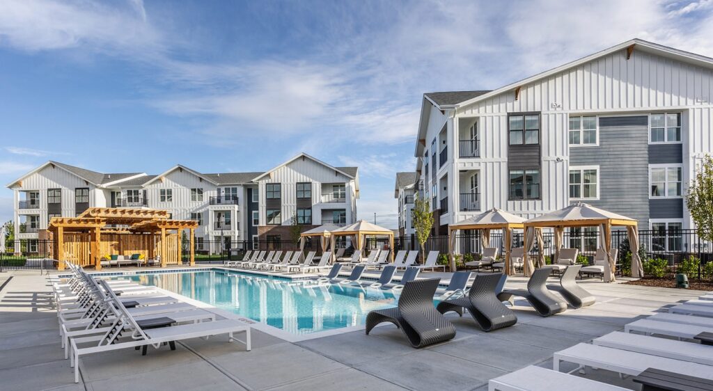Modern apartment complex with a large outdoor swimming pool, surrounded by white lounge chairs and shaded cabanas. A wooden pergola sits to the left, and the buildings feature gray and white facades under a blue sky.