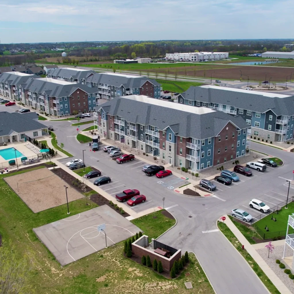 Aerial view of a modern apartment complex with blue-gray buildings, parking lots, and a playground/basketball court nearby surrounded by green lawns.