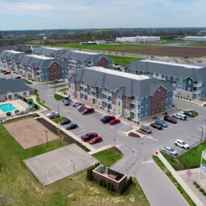 Aerial view of a modern apartment complex with blue-gray buildings, parking lots, and a playground/basketball court nearby surrounded by green lawns.