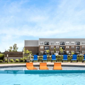 Poolside scene with orange lounge chairs by a bright blue pool, blue chairs lined up behind a black fence and apartment buildings in the background.