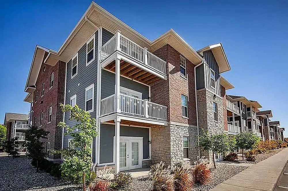 Modern multi-story apartment building with brick and stone facade and white balconies along a sidewalk.