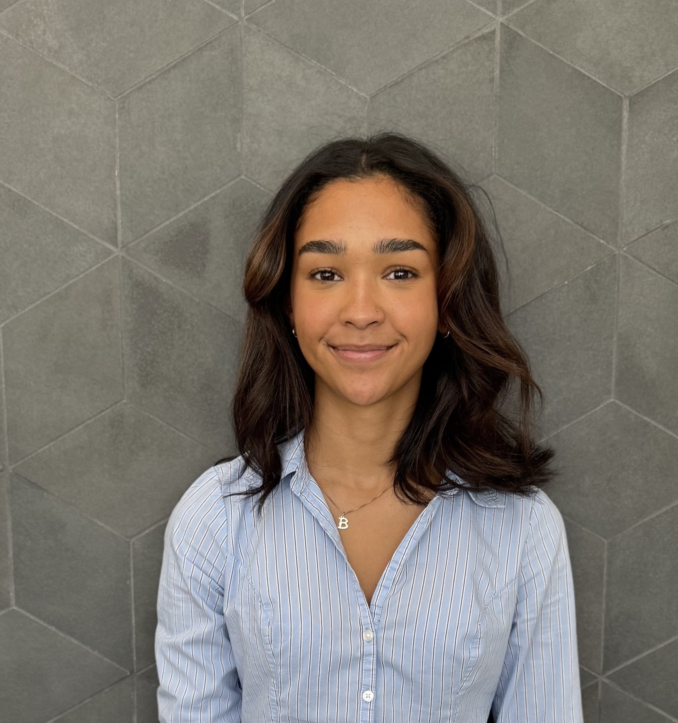 A young woman with wavy dark hair, wearing a light blue striped shirt and a B necklace, smiles in front of a gray geometric patterned wall.